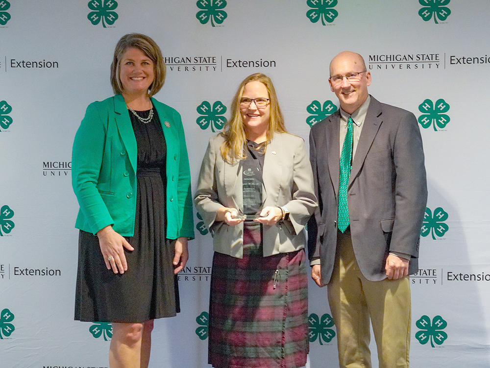 Three individuals at a backdrop. The center person holds an award.